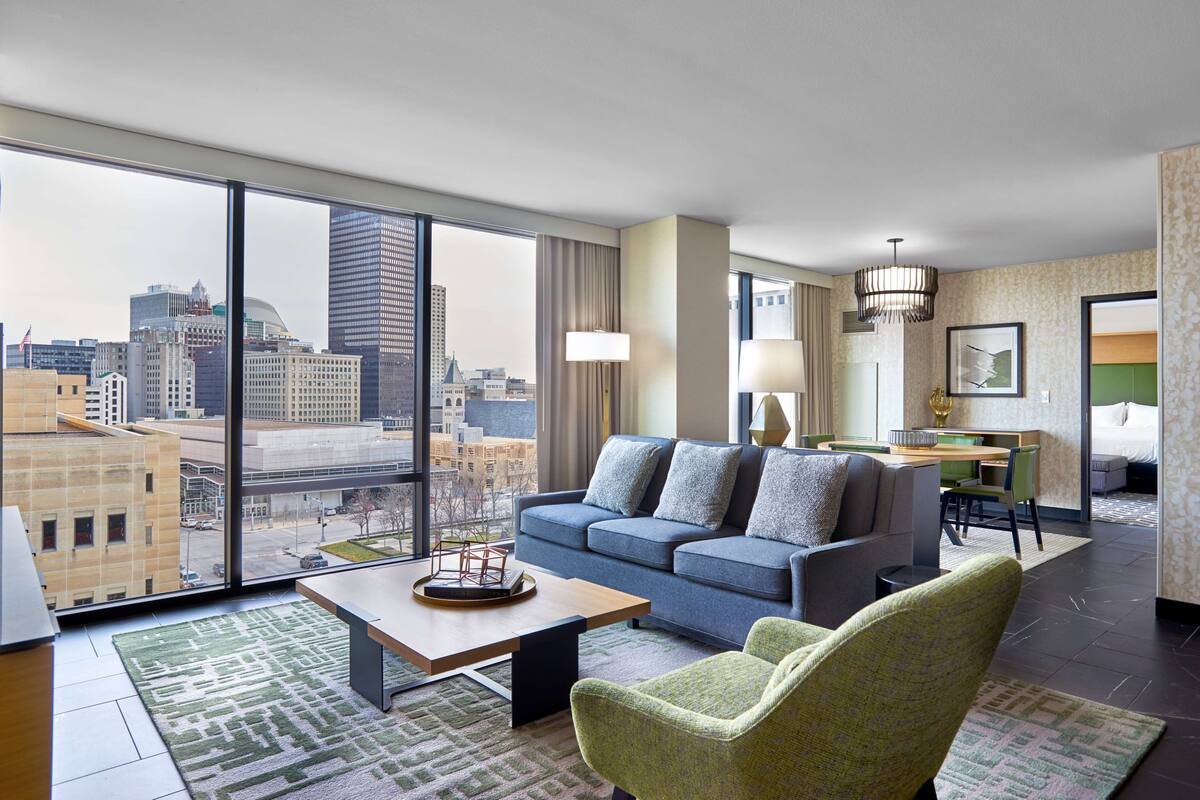 A modern hotel room with a large city view, a sofa, a dining area, and a small table with a decorative item.