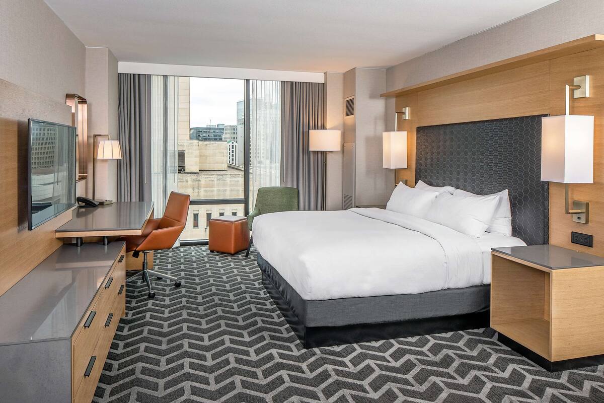 A hotel room with a large bed, a desk with a chair, a TV, and a view of the city through large windows.