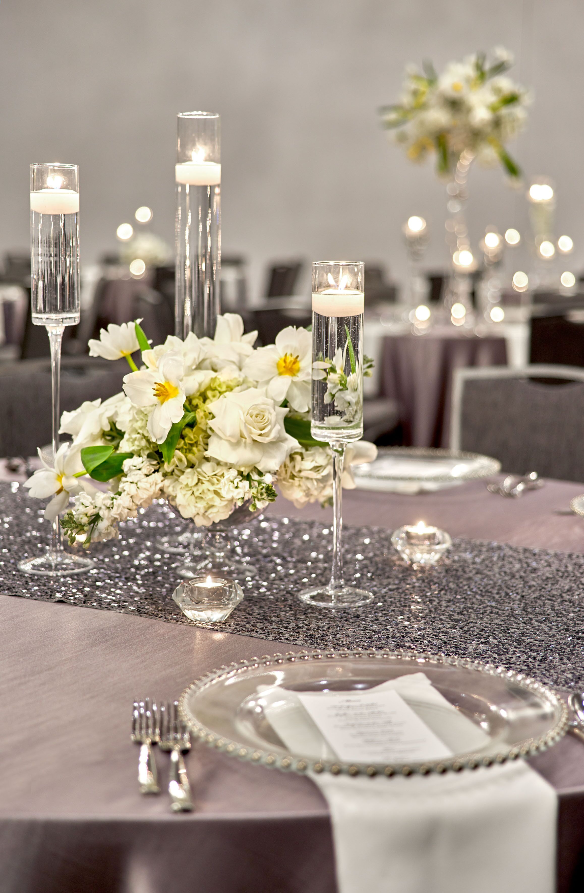 A table set with white flowers, candles, and silverware, suggesting a formal dining setting.