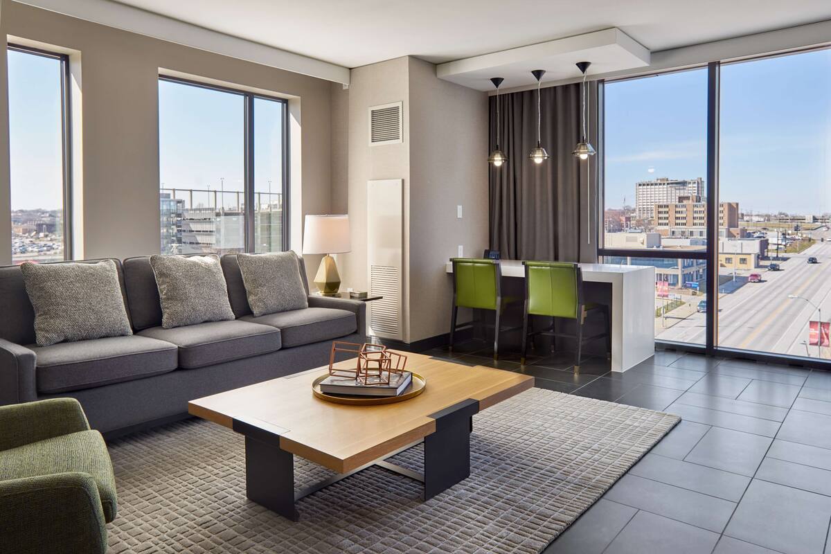 A modern hotel room with a large window, a sofa, a coffee table, and a dining area with chairs.