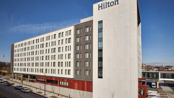 A modern hotel building with a prominent 'Hilton' sign, multiple floors, and a parking area with a bus stop.