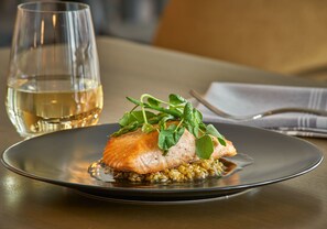 A plate of food with a fish fillet, greens, and grains, accompanied by a glass of white wine on a table. - Hilton Des Moines Downtown (Des Moines)