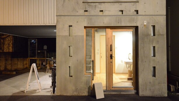 Un edificio moderno con fachada de hormigón, puerta de madera y un interior visible con un lavabo y encimera.