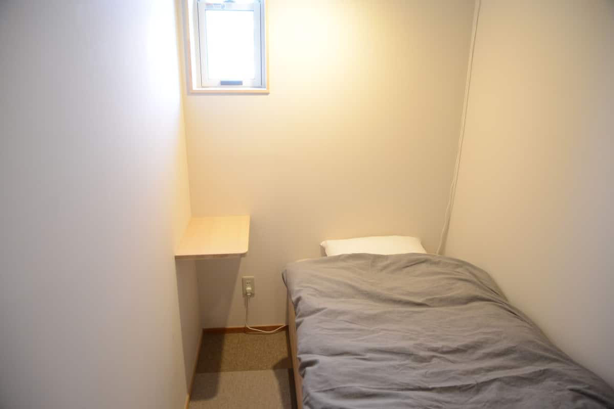 A small, narrow room with a single bed, a window, and a wall-mounted shelf.