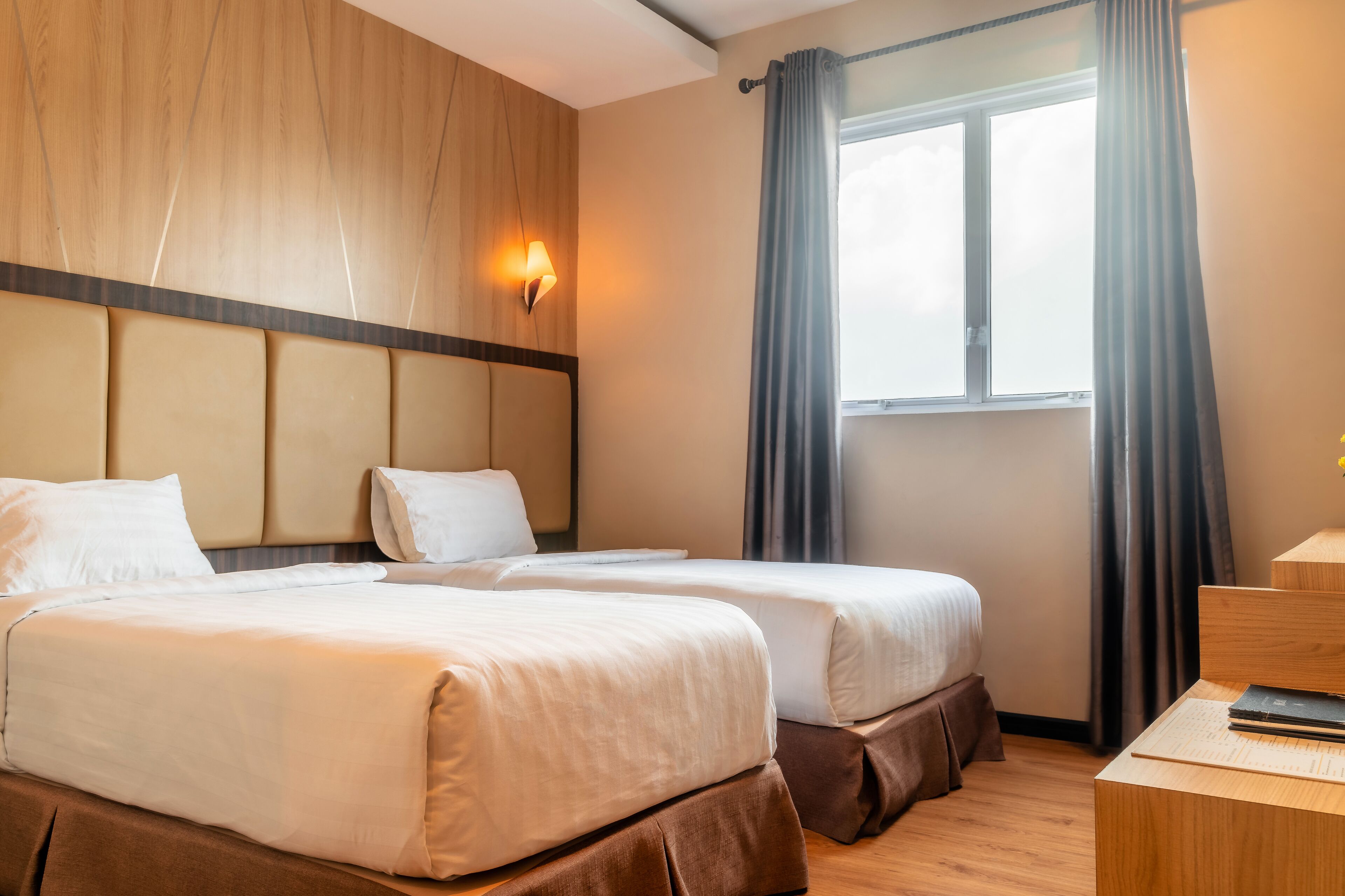 A hotel room with two beds, a wooden headboard, a nightstand, and a window with curtains.