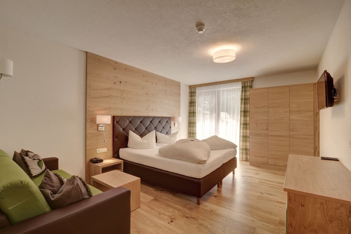 A modern bedroom with a wooden headboard, a bed with white linens, a green sofa, a wooden nightstand, and a wooden wardrobe.