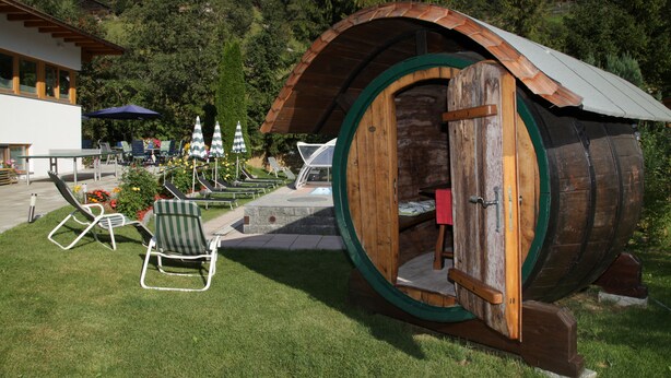 Une structure rustique en bois, en forme de tonneau, avec un toit incurvé, une petite table et une chaise à l’extérieur.