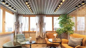 Une pièce chaleureuse avec un fauteuil suspendu, un canapé et une petite table. De grandes fenêtres sont ornées de rideaux, et on y trouve également un radiateur et une plante en pot.