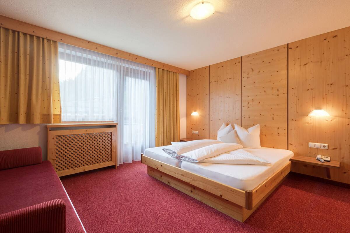 A hotel room with a wooden bed, a red sofa, a window with curtains, and a wall-mounted lamp.