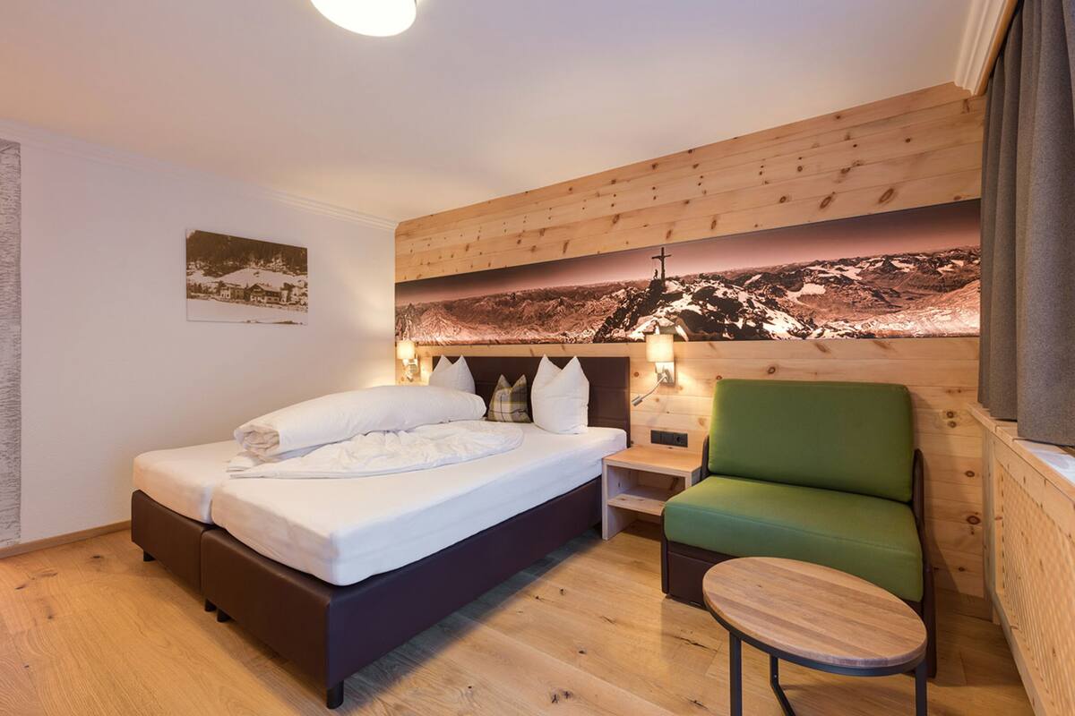 A hotel room with a wooden bed, a green armchair, a small wooden table, and a wall-mounted lamp.