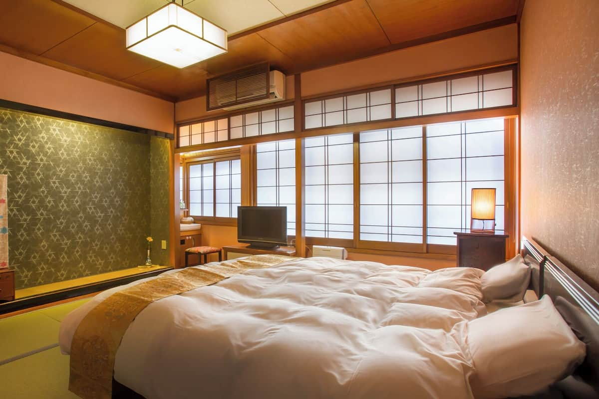 A traditional Japanese-style room with tatami flooring, a bed with white bedding, a television, and a window with shoji screens.