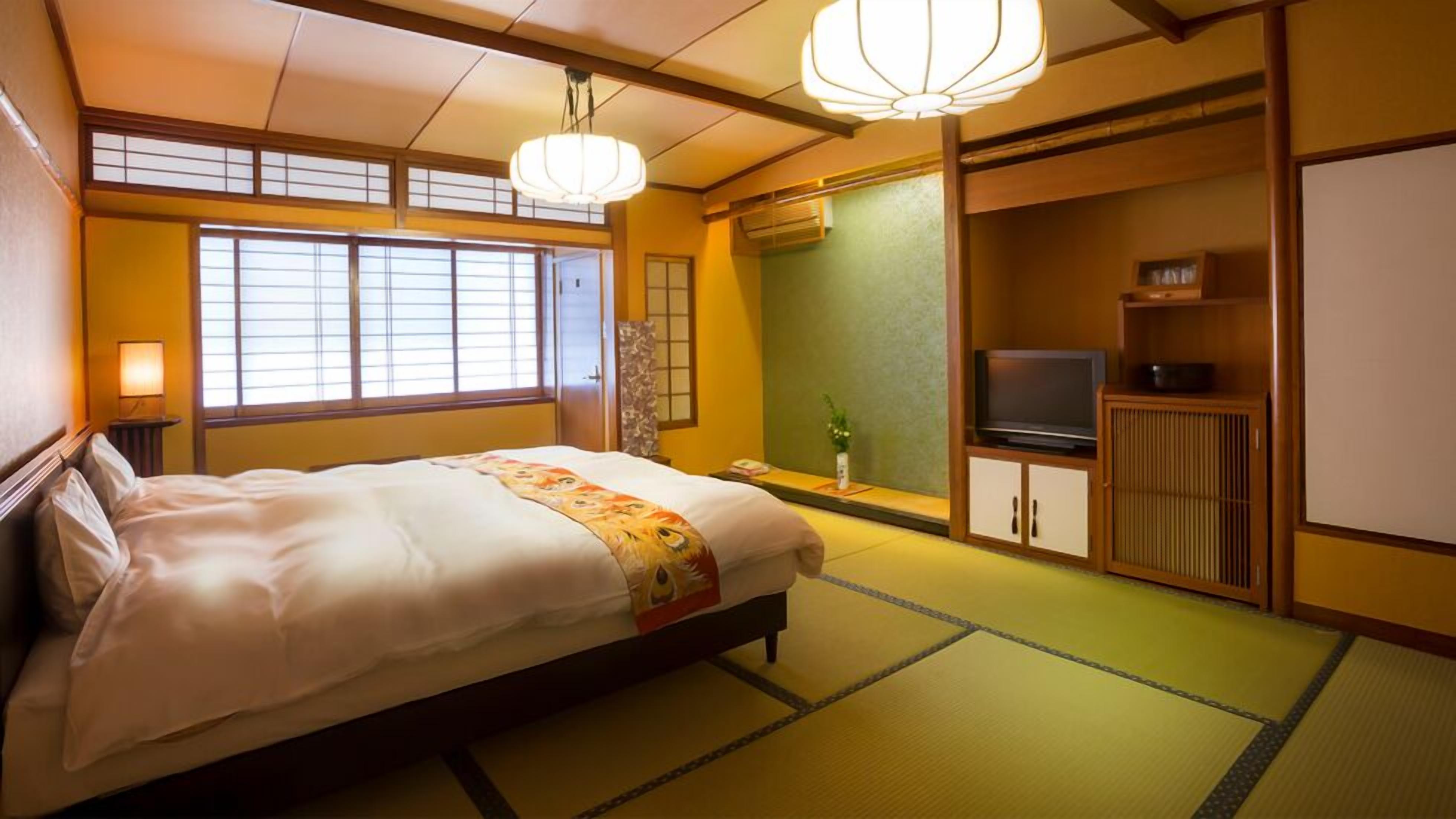 Een traditionele Japanse kamer met tatami vloeren, een bed met een bloemenprint dekbed, een televisie op een houten kast en een shoji scherm.