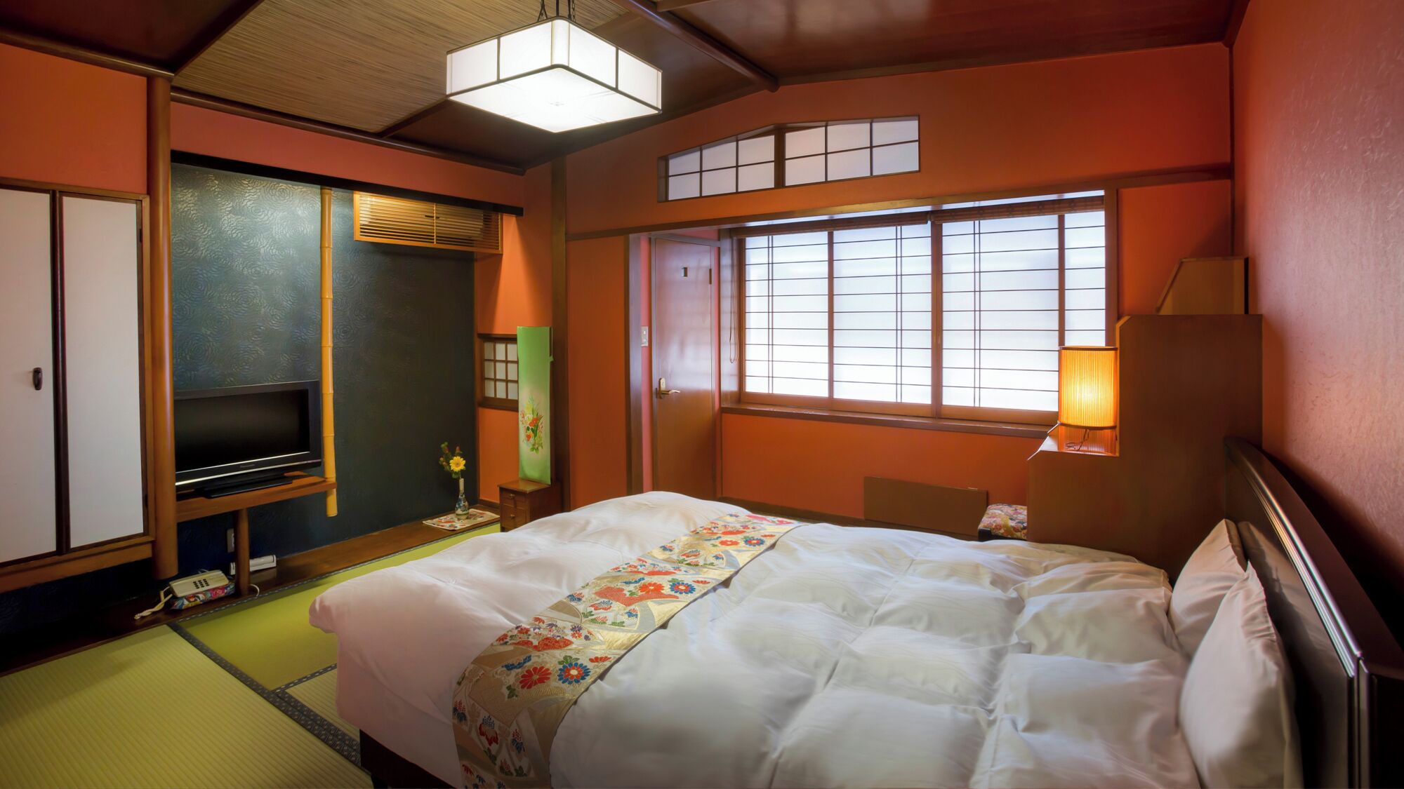 A traditional Japanese-style hotel room with tatami flooring, a bed with a floral bedspread, a television, and sliding doors.