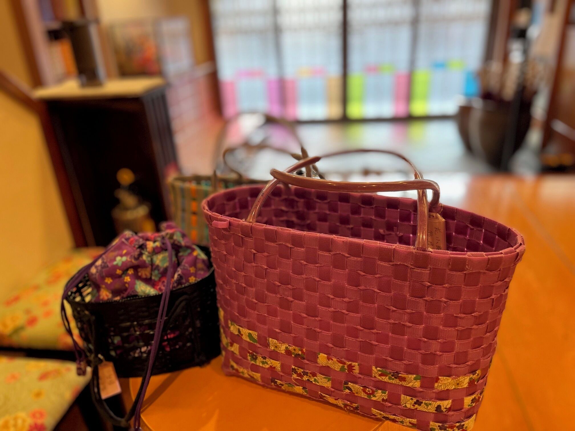 A wicker basket with a handle, a black woven bag, and a floral-patterned bag on a wooden surface.