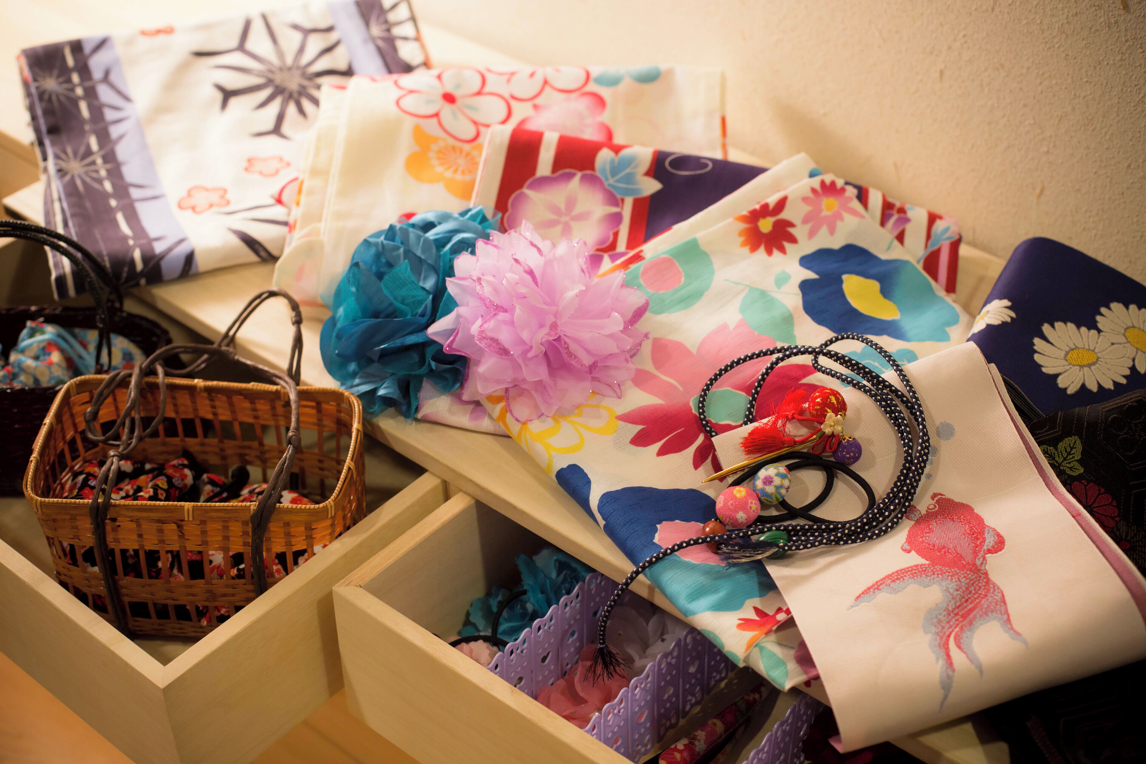 Un espacio de almacenamiento con una cesta de mimbre que contiene ropa doblada, un cajón con ropa doblada y un estante con diversos objetos, incluyendo un collar de cuentas y un tejido con estampado floral.