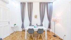 A room with a white table, two chairs, a small white side table, a floor lamp, and grey curtains. - Dominik Apartment (Budapest)