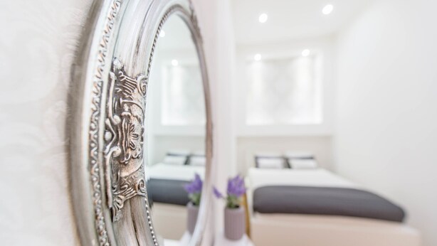 A bedroom with a large ornate mirror, a bed with gray bedding, and a small potted plant. - Dominik Apartment (Budapest)