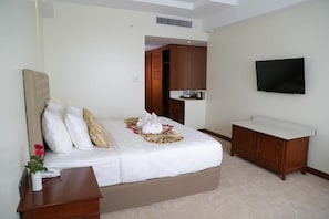 A hotel room with a bed, a bedside table with a vase of flowers, a flat-screen TV mounted on the wall, and a wooden cabinet. - Pacific Hotel (Yangon)