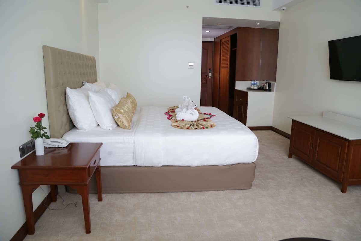 Habitación de hotel con una cama grande, una mesita de noche de madera, un televisor montado en la pared y una kitchenette al fondo.