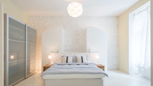 A modern bedroom with a white brick wall, a large bed with a striped bedspread, a bedside table with a lamp, and a hanging light fixture.