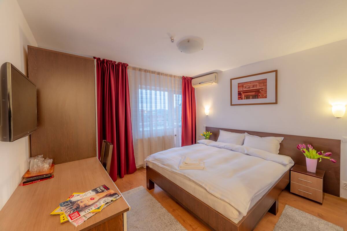 A hotel room with a bed, bedside table, TV, and a window with curtains.