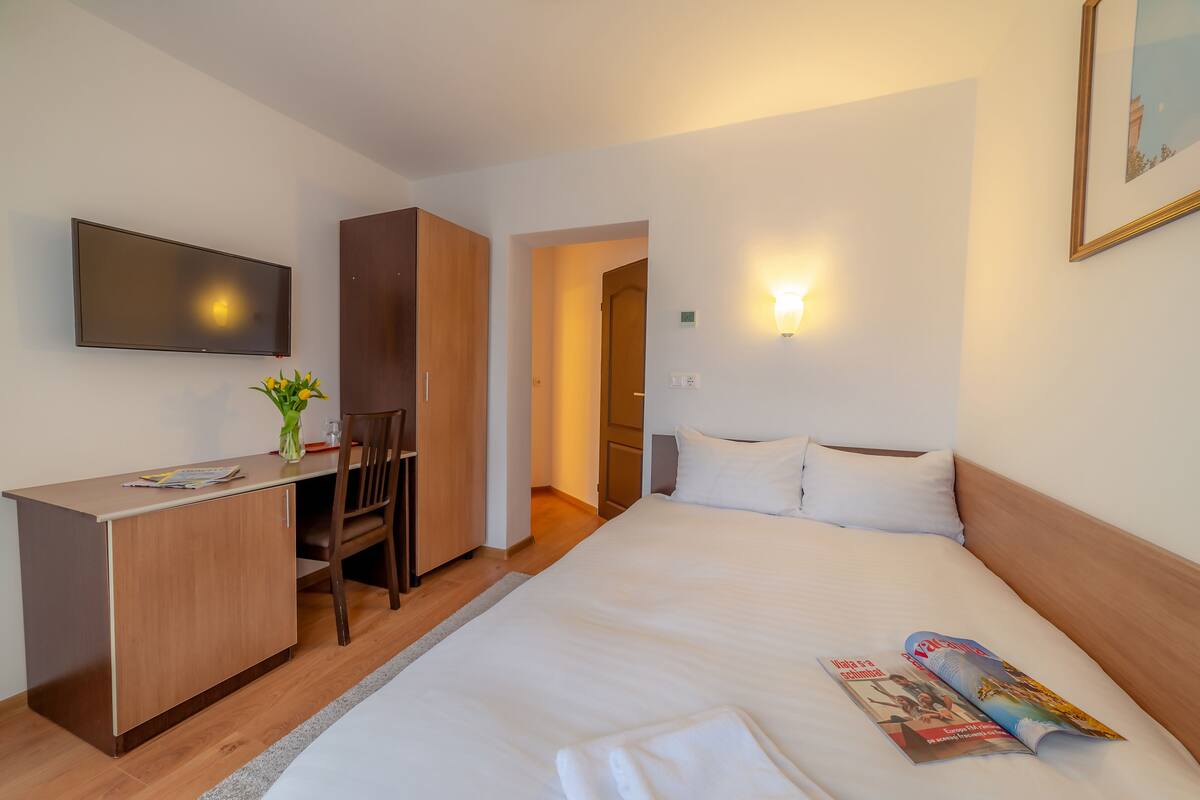 A hotel room with a bed, a desk, a chair, a television, and a wall-mounted light fixture.
