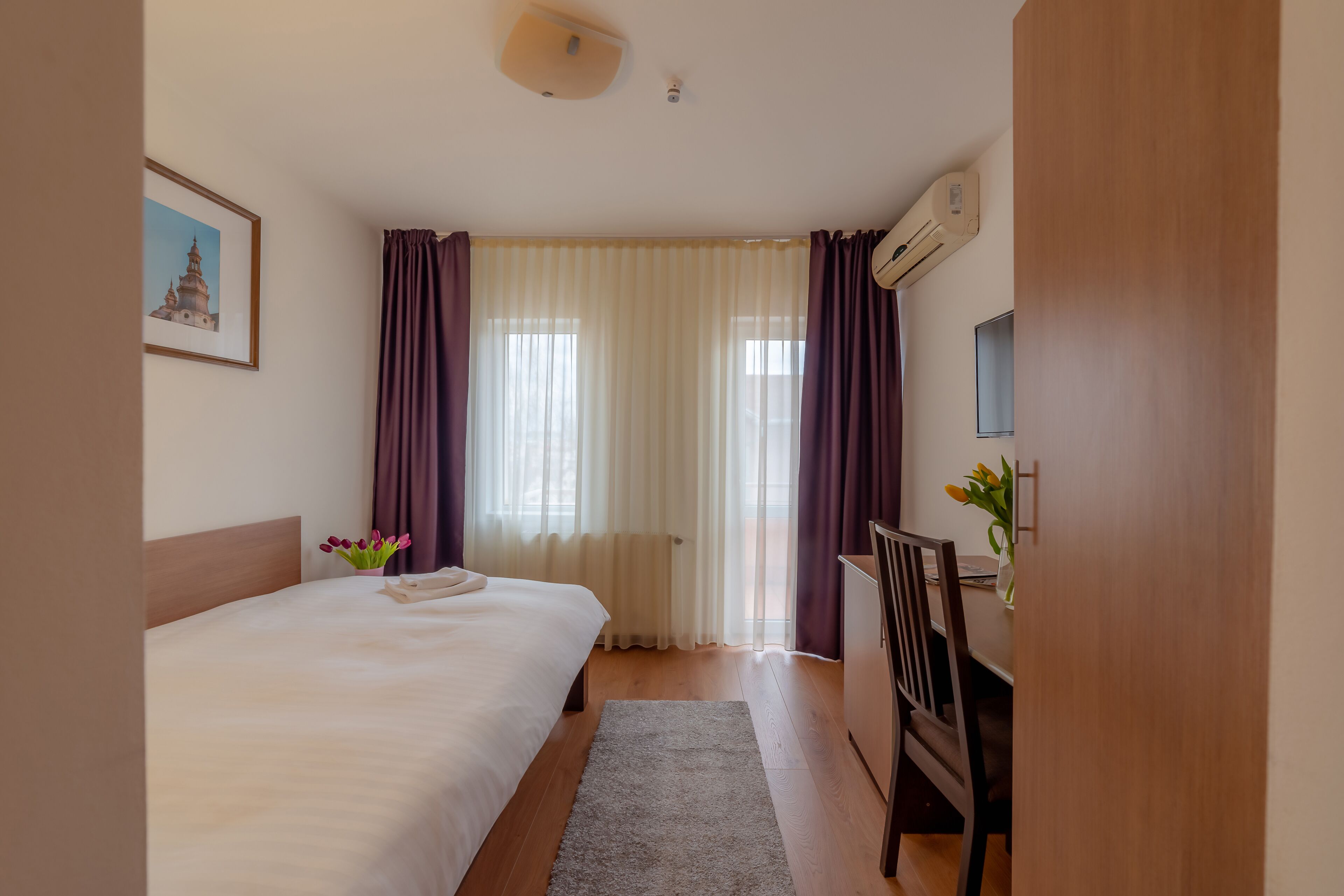 Et hotelværelse med seng, skrivebord, stol, et vindue med gardiner, en radiator og en aircondition.
