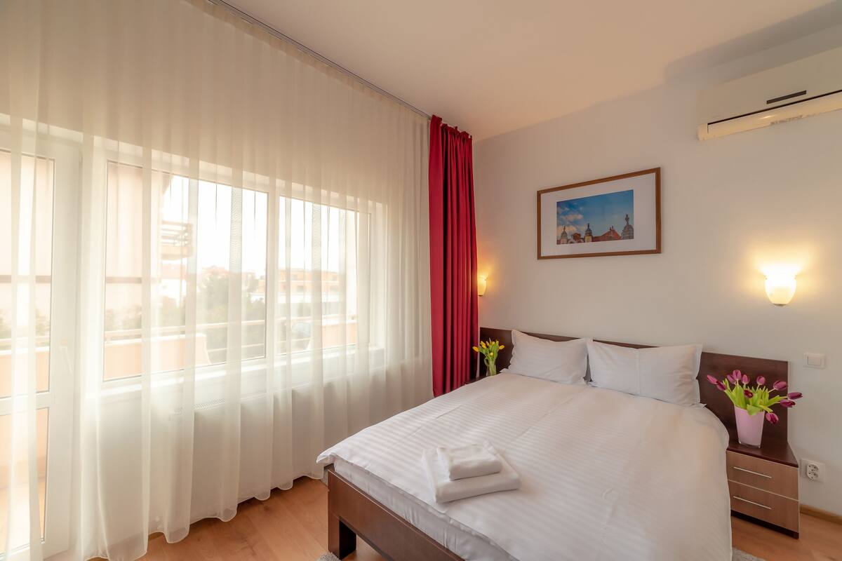 A hotel room with a large bed, a nightstand with a vase of flowers, a painting on the wall, and a window with sheer curtains.