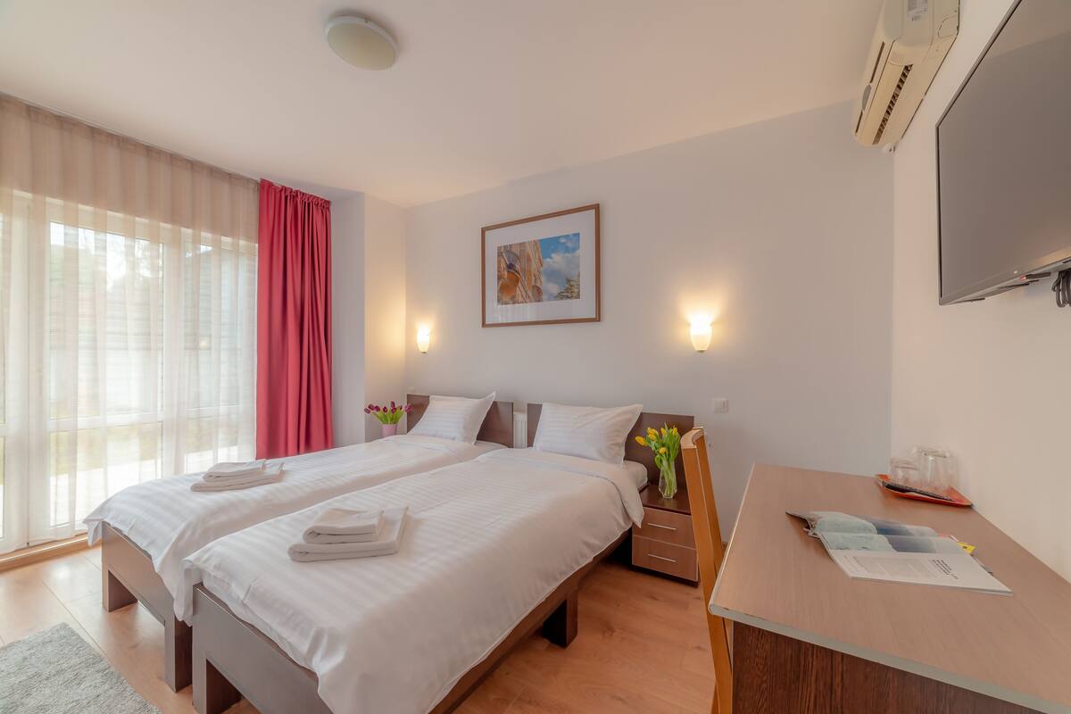 A hotel room with two beds, a desk, a TV, and a window with curtains.