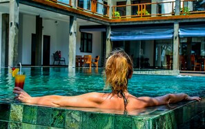 A person relaxing in a pool with a drink, surrounded by a building with outdoor seating and a covered area. - Hikka FnR Villa (Hikkaduwa)