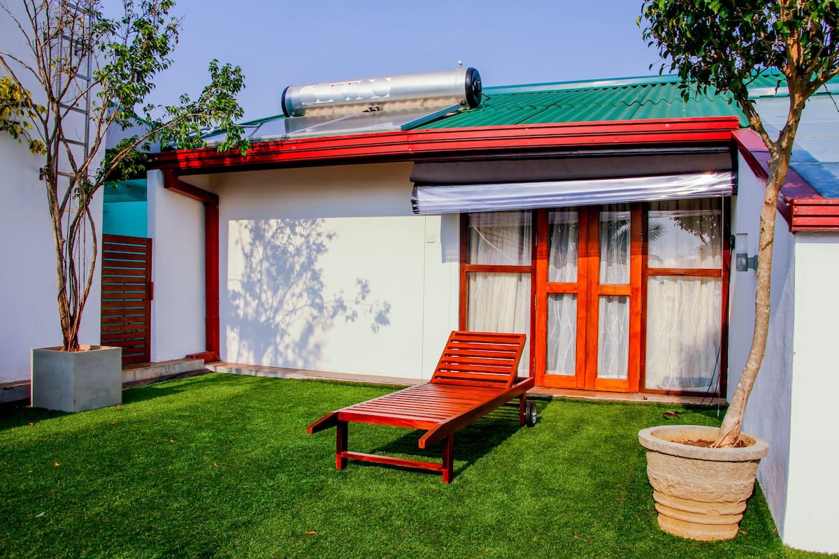 A red lounge chair on a well-maintained lawn, a large potted tree, and a building with a green roof and red accents.