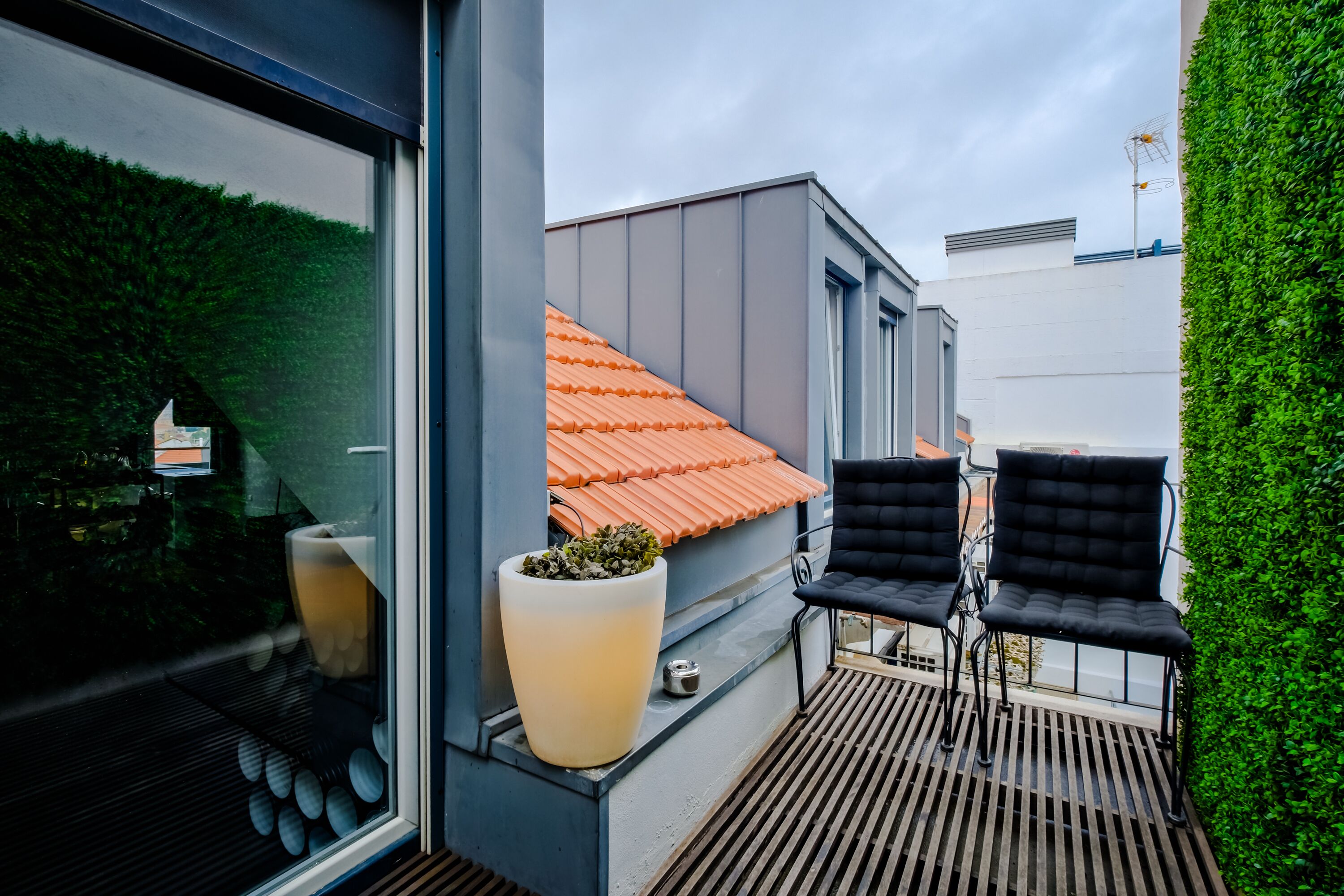 A modern outdoor balcony with a potted plant, two black chairs, and a red-tiled roof visible through the window.