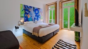A modern bedroom with a large bed, a painting on the wall, and a view of greenery through the window. - Chiado Arty flats (Lisbon)