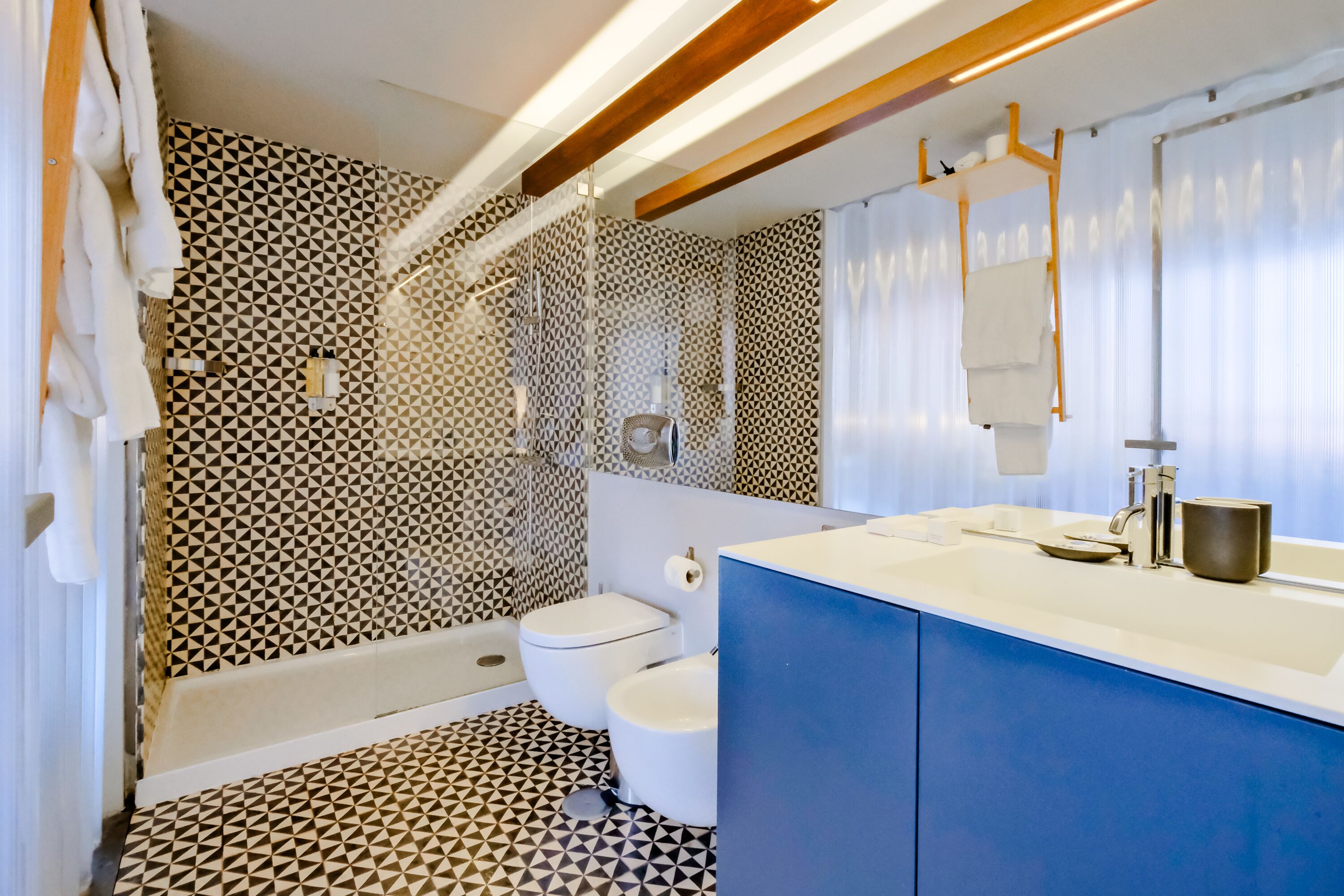 A modern bathroom with a geometric patterned floor, a white toilet, a bathtub, and a blue vanity with a sink and mirror.