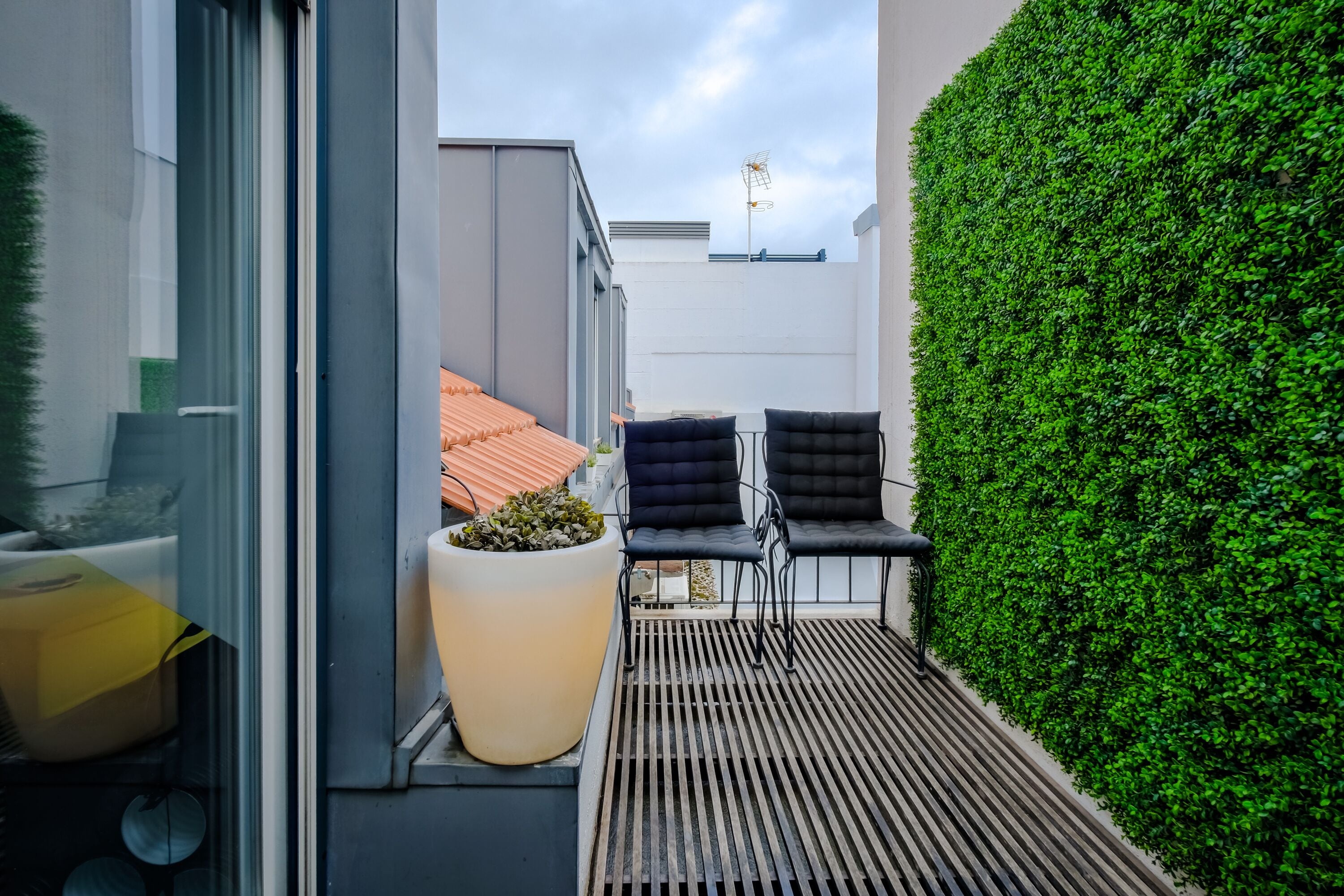 A modern outdoor seating area with two black chairs, a large planter with greenery, and a wooden slatted floor.
