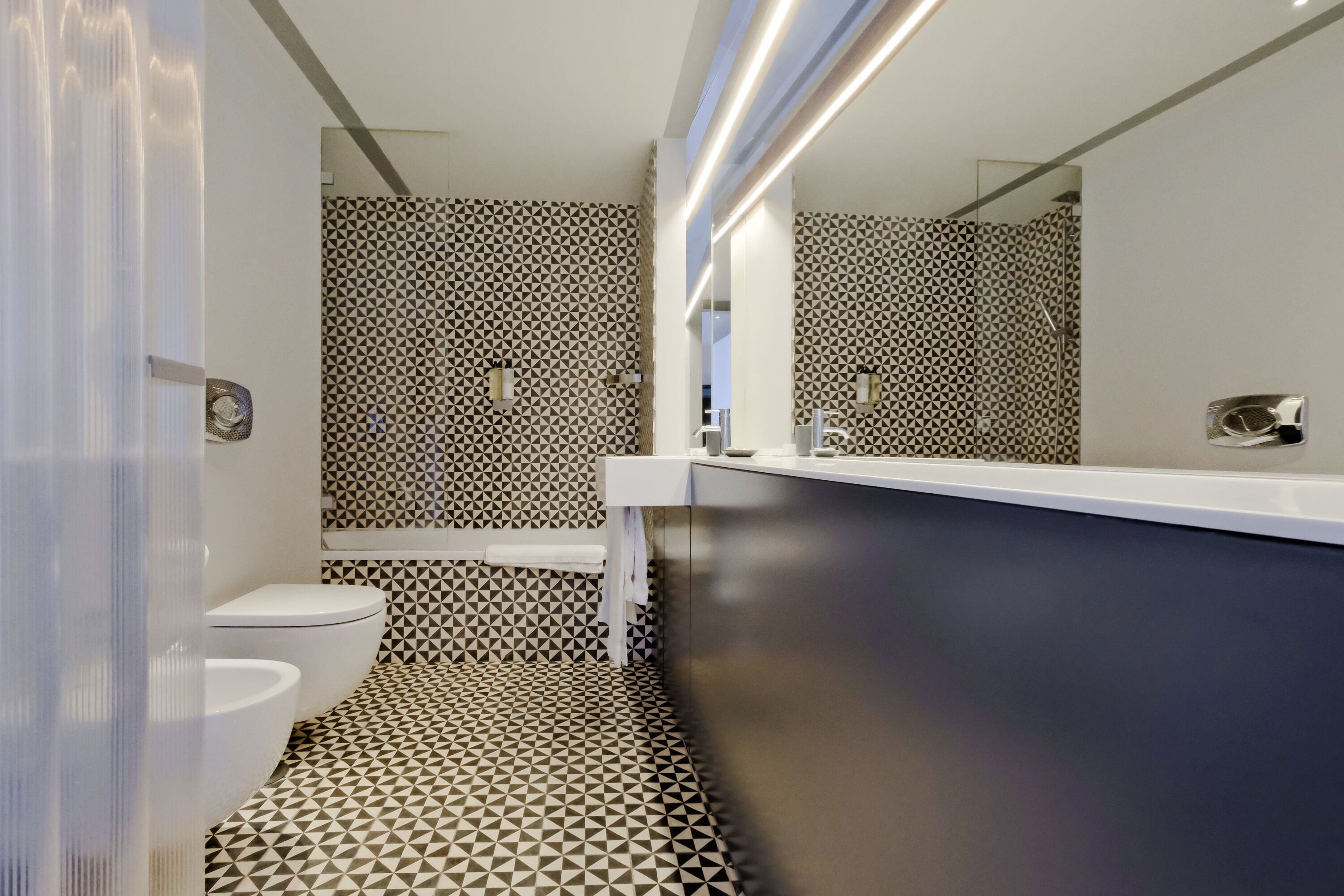 A modern bathroom with a geometric patterned floor, a white toilet, and a contemporary sink area with a mirror.