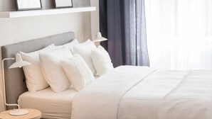 Ein ordentlich bezogenes Bett mit weißer Bettwäsche und einem grauen Kopfteil, eine Nachttischlampe und ein kleiner runder Beistelltisch mit einer Lampe.