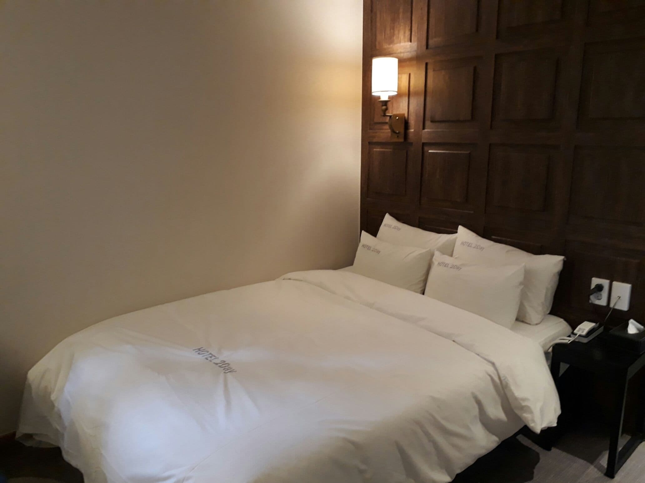Una cama bien hecha con ropa de cama blanca y almohadas que dicen "HOTEL 20BY", en una habitación con paneles de madera en la pared y una mesita de noche con una lámpara y un teléfono.