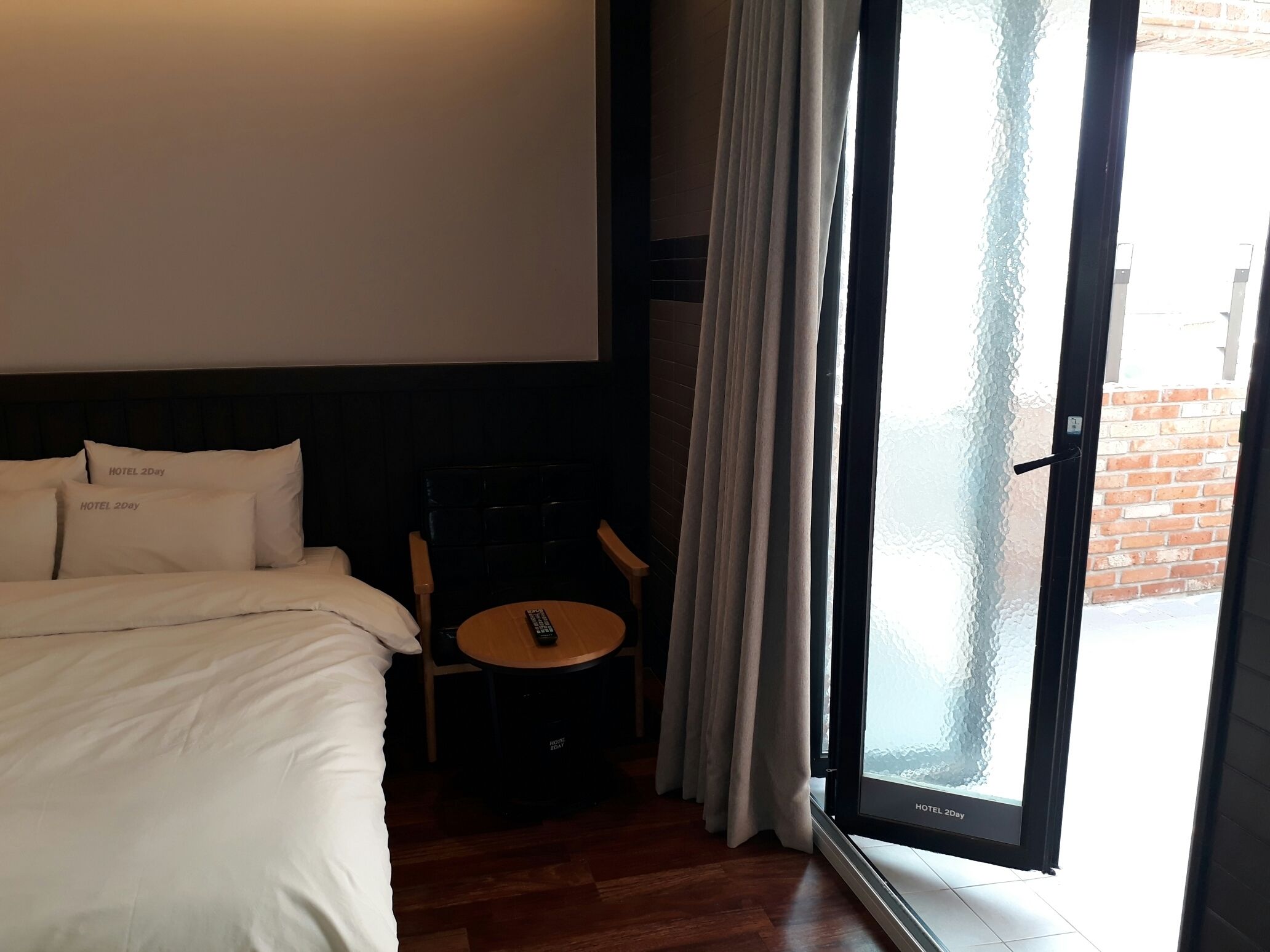 Une chambre d’hôtel avec un lit, une petite table sur laquelle se trouve une télécommande, et une vue sur l’extérieur à travers une porte vitrée.