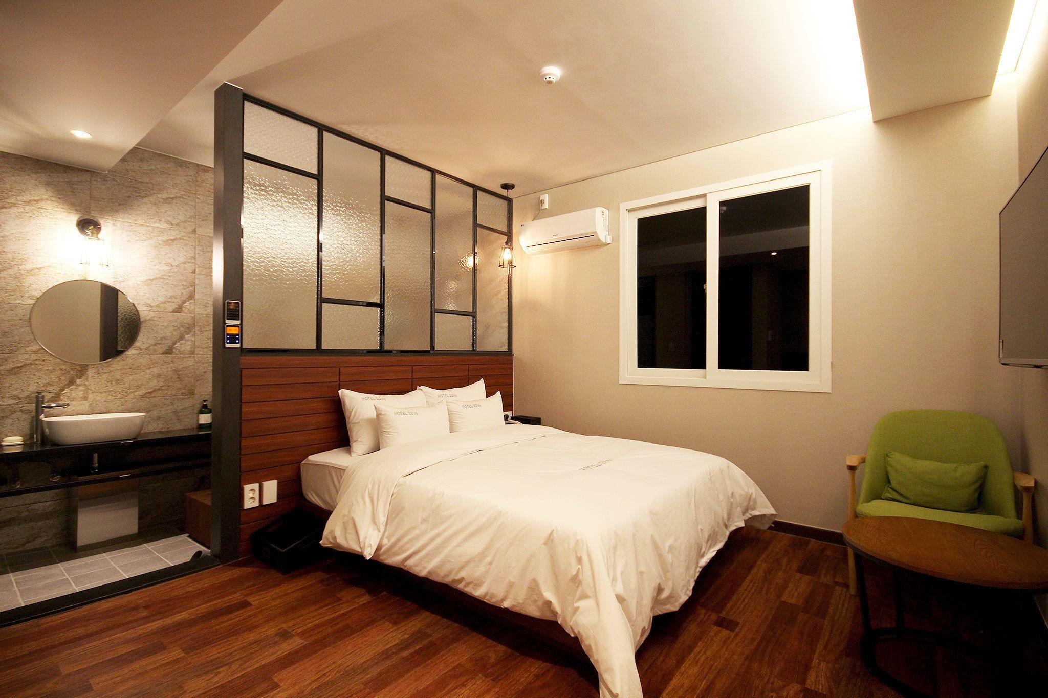 A hotel room with a wooden headboard, a large bed, a green chair, a round mirror, a bathroom with a sink, and a window with blinds.
