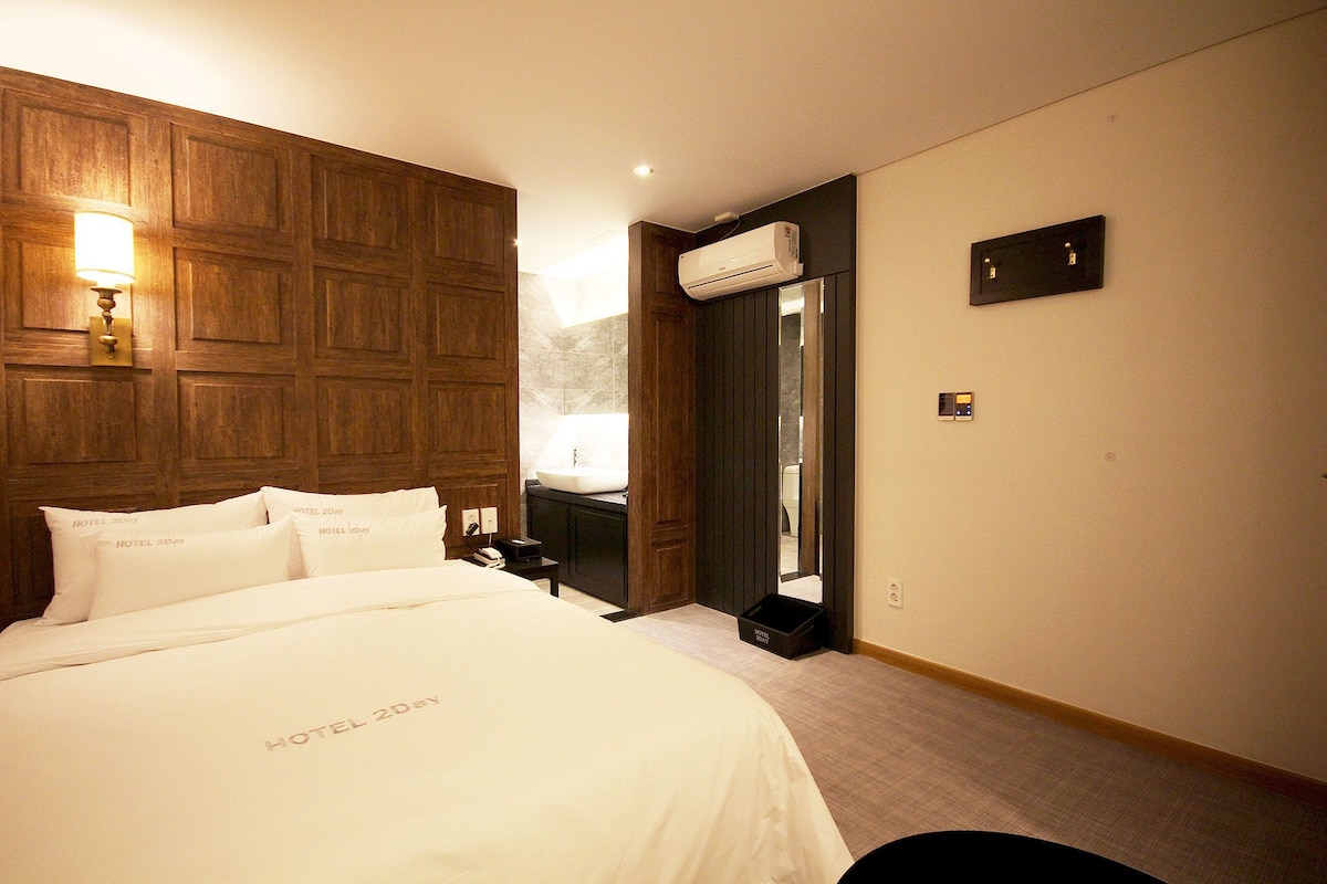 A hotel room with a large bed, a nightstand, a wall-mounted air conditioner, a mirror, and a door leading to a bathroom.