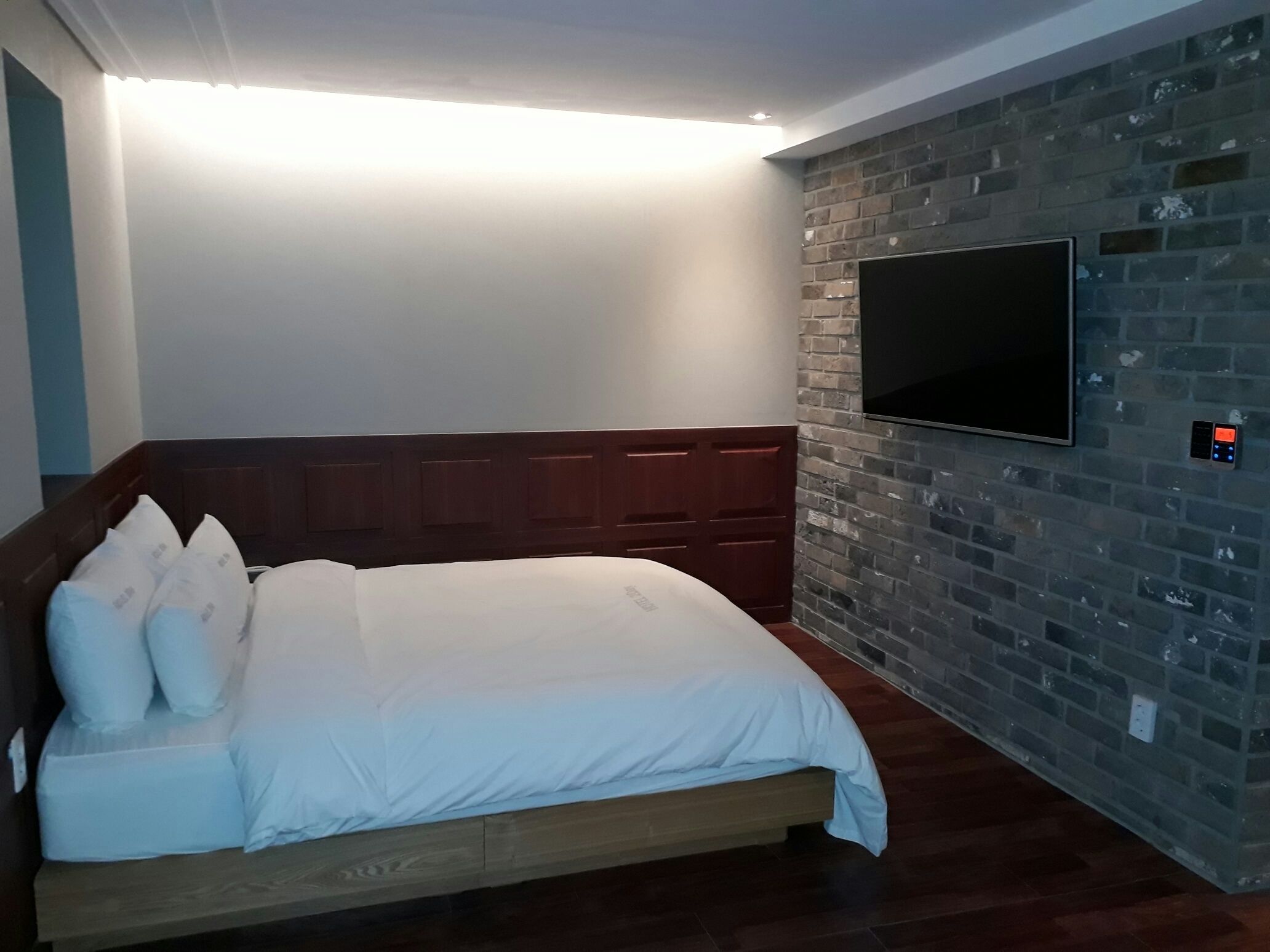 A bedroom with a wooden headboard, a large bed with white bedding, a flat-screen TV mounted on a brick wall, and a control panel for the TV.