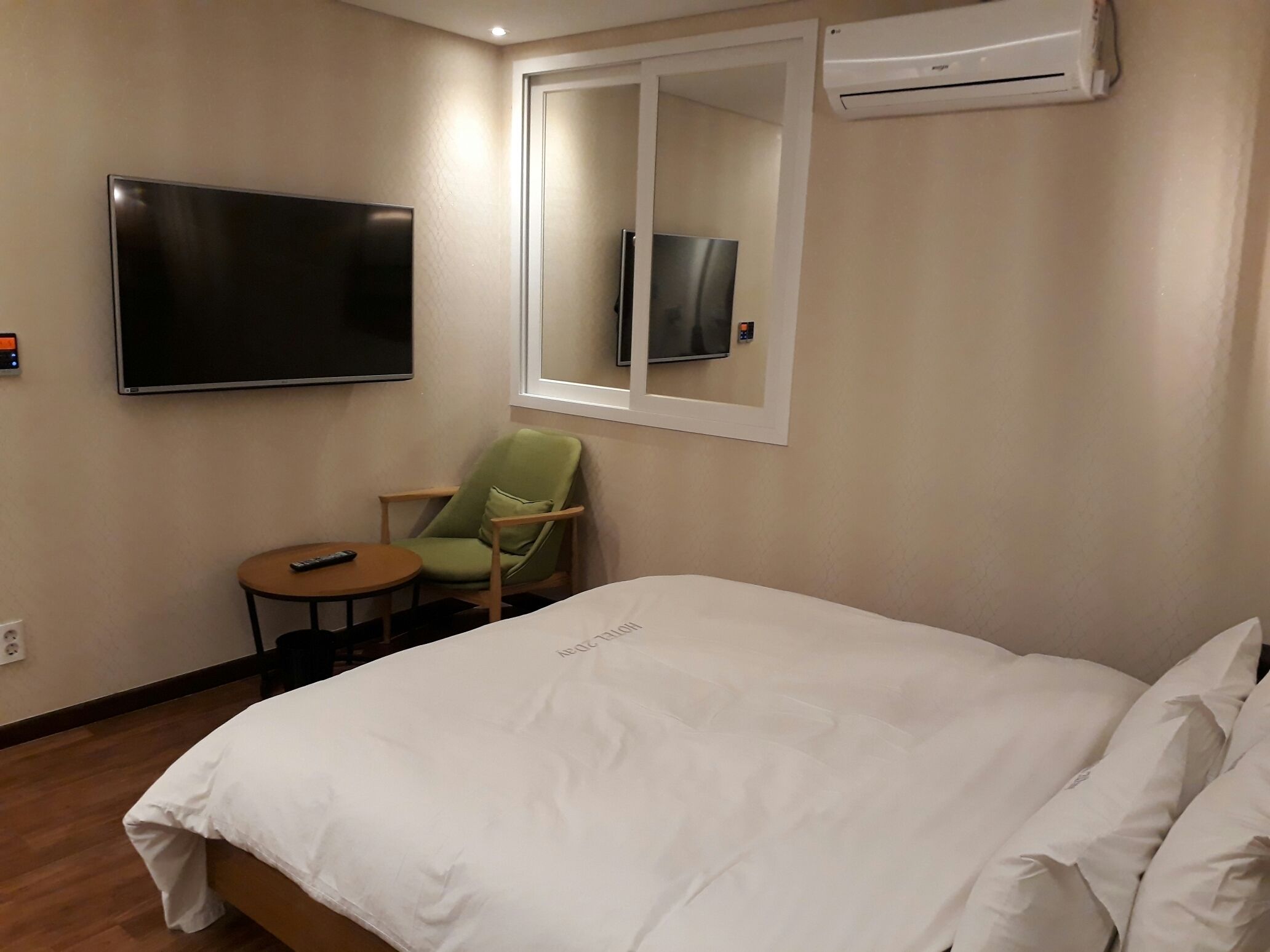 A hotel room with a large bed, a green armchair, a small round table, a TV mounted on the wall, and an air conditioning unit.