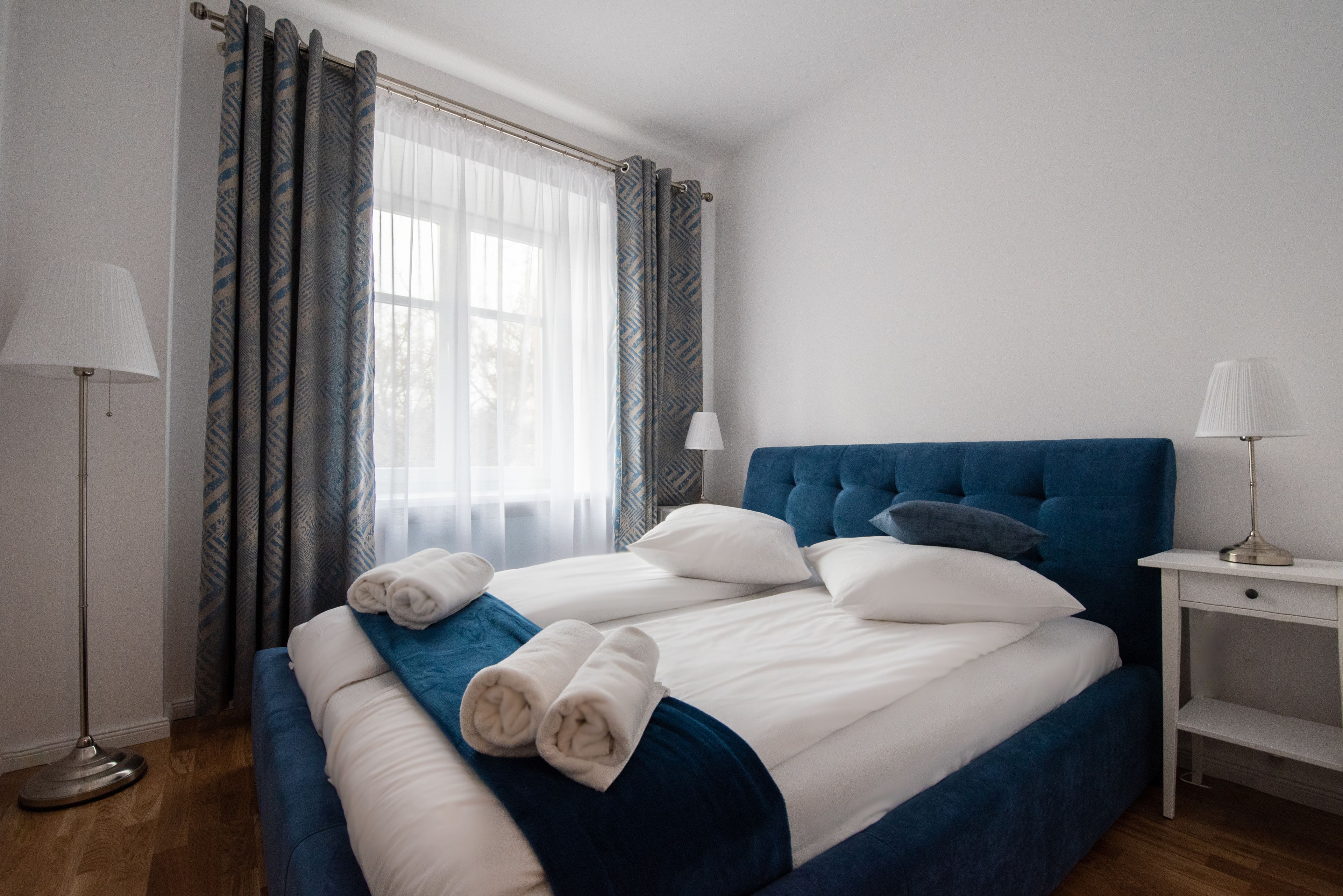 A neatly made bed with white and blue bedding, two bedside lamps, and a window with patterned curtains.