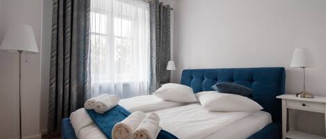 A neatly made bed with white and blue bedding, two bedside lamps, and a window with patterned curtains.