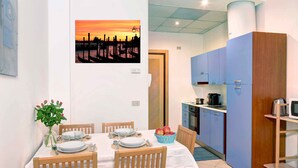 Un coin repas avec une table dressée pour quatre personnes, un vase rempli de fleurs et un tableau représentant un coucher de soleil.