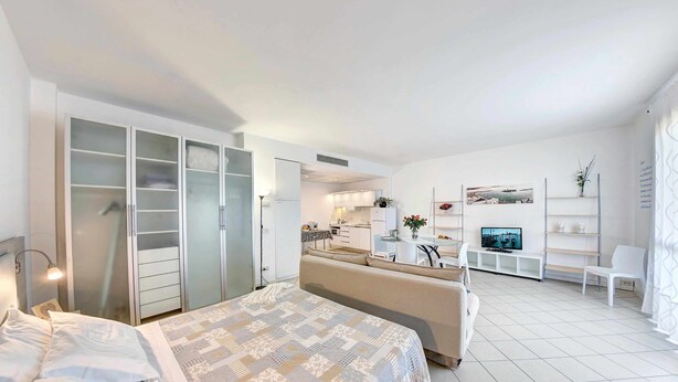 Une chambre à coucher moderne et bien éclairée, avec un grand lit, une armoire et un coin cuisine en arrière-plan.