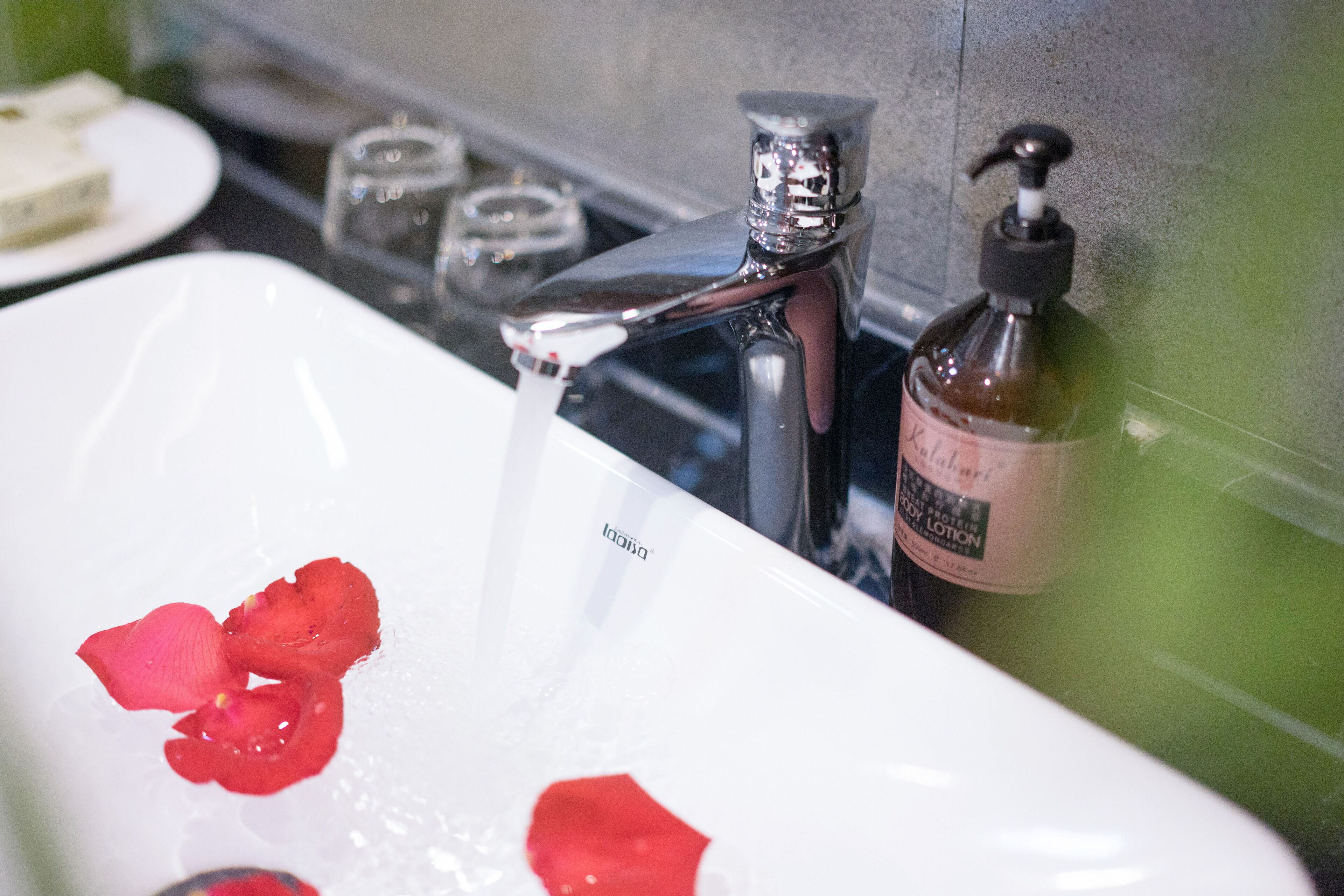 Un lavabo de salle de bain avec un robinet chromé, un flacon de lotion pour le corps et des pétales de rose flottant dans l’eau.