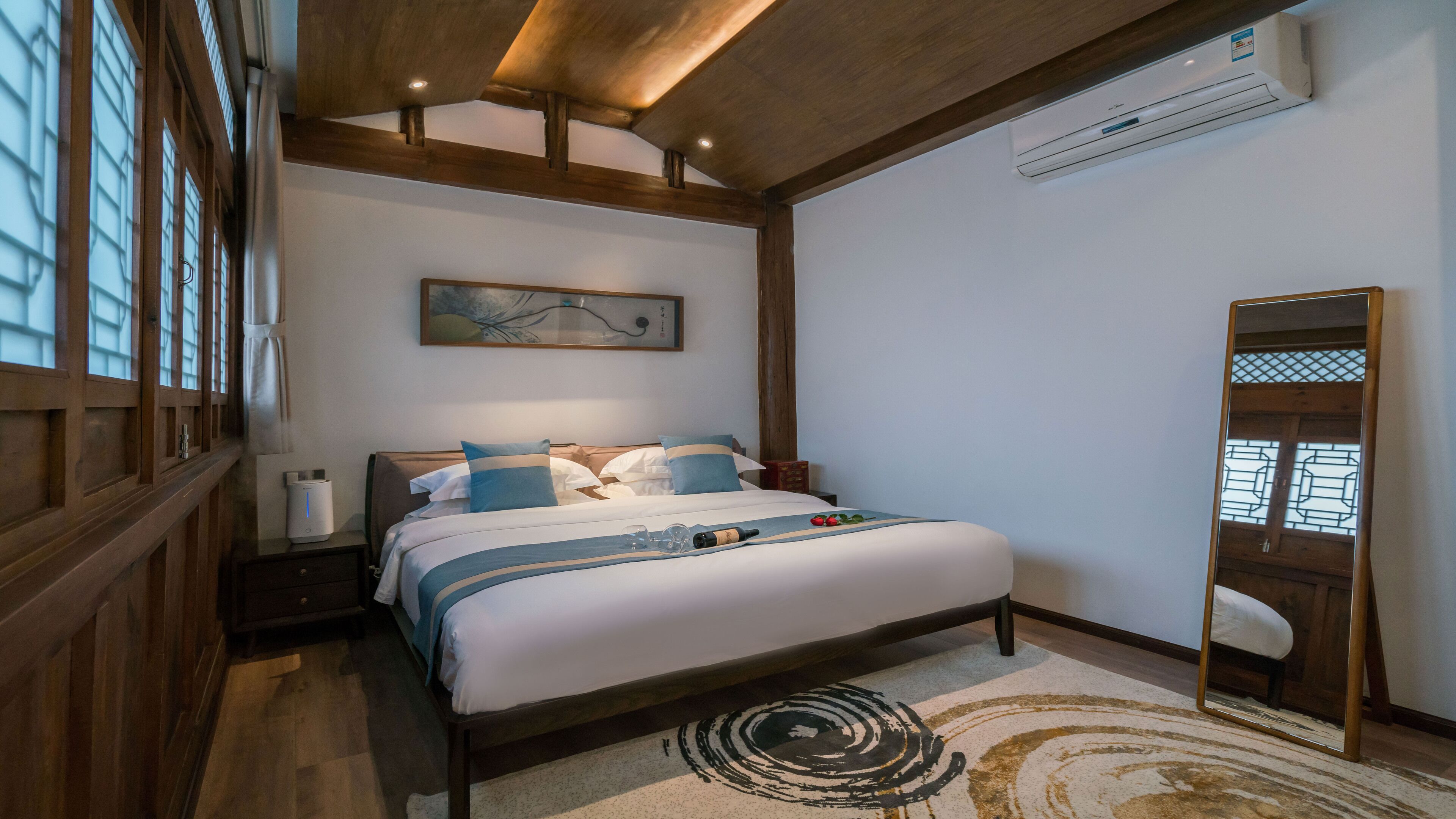 A bedroom with a large bed, wooden beams, an air conditioner, a painting, and a mirror.