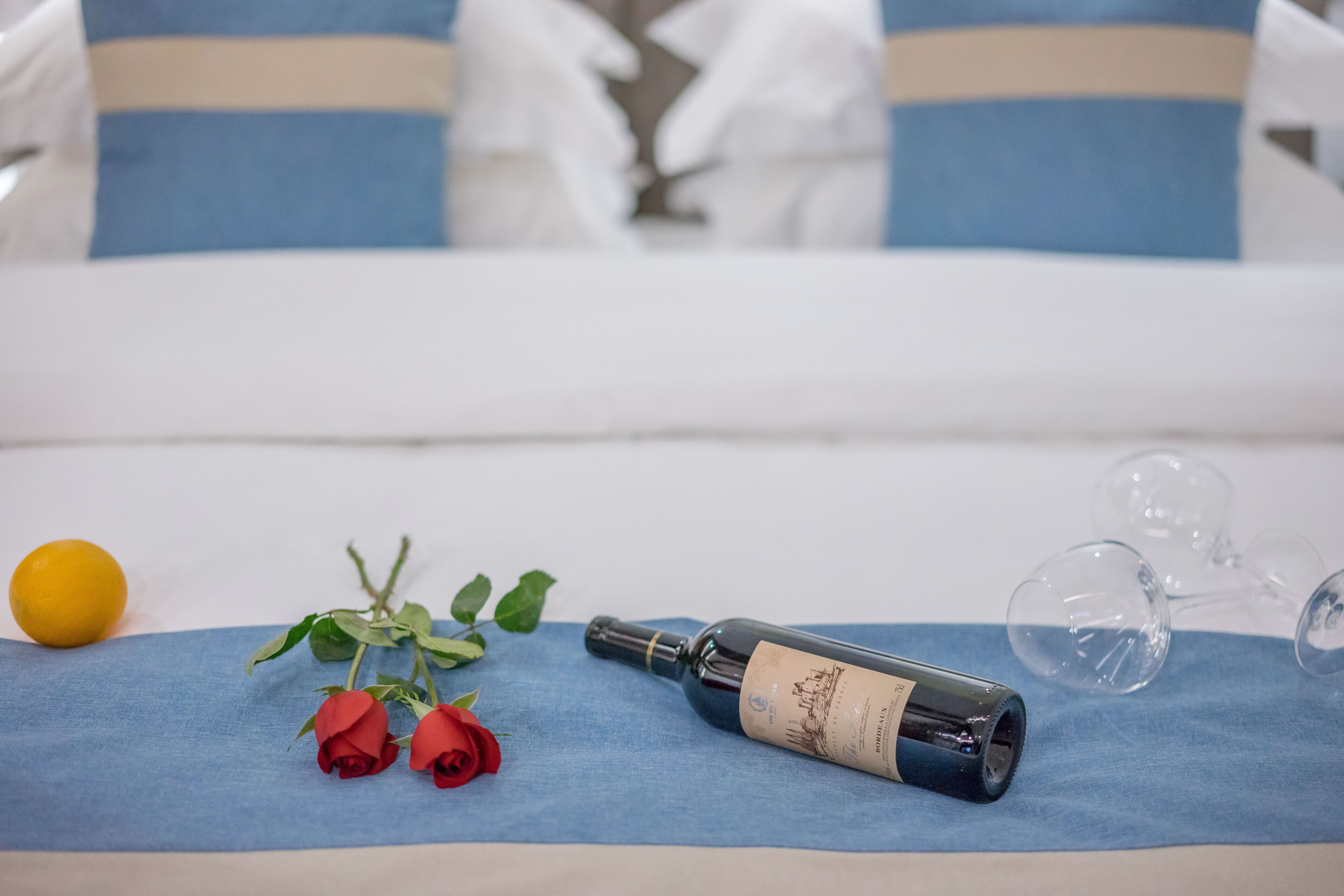 A bed with a blue and white striped pillow, a bottle of wine, two red roses, a lemon, and wine glasses on a blue cloth.
